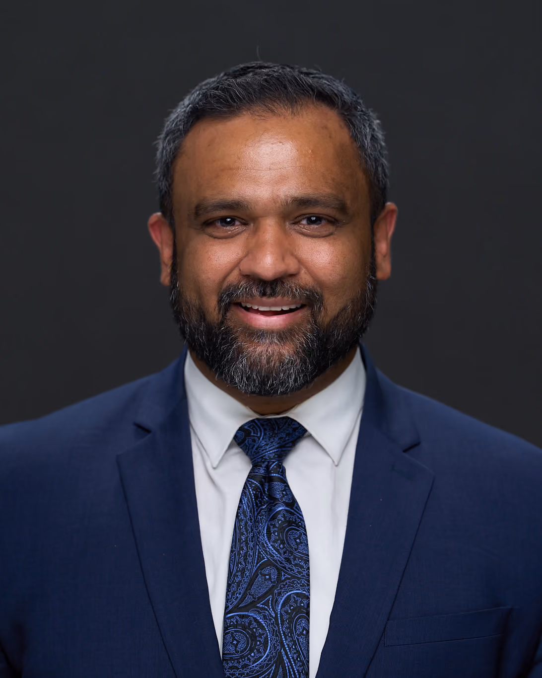 Neel Patel, M.D., board-certified hip and knee replacement surgeon, photographed in a navy suit against a charcoal studio background